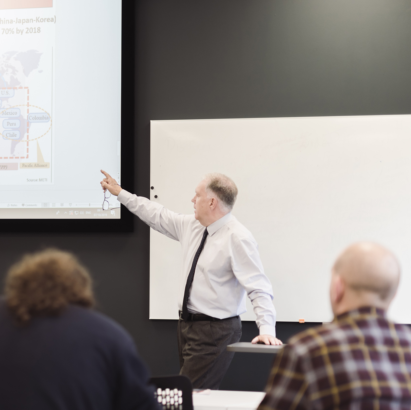 Students in a lecture - the lecturer is pointing at the projection of the presentation slides, while students take notes